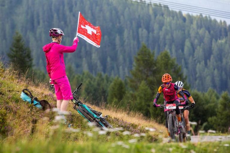 Someone holding a Swiss flag while two bikers head past