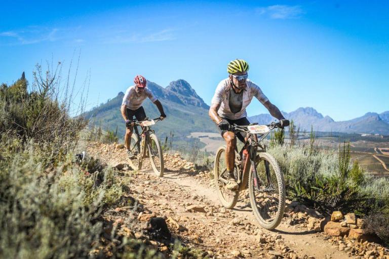 Two bikers heading through a rocky path with mountains in the background