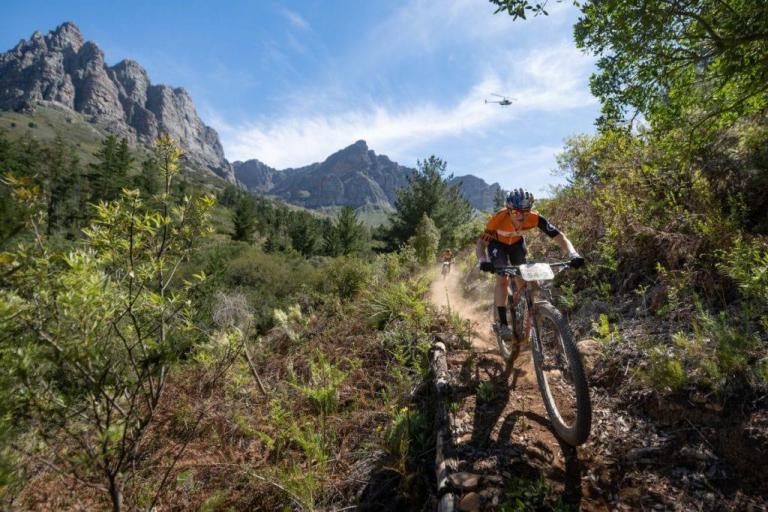 An incredible shot of the bikers on a small path with hills in the background and a helicopter above one biker's head
