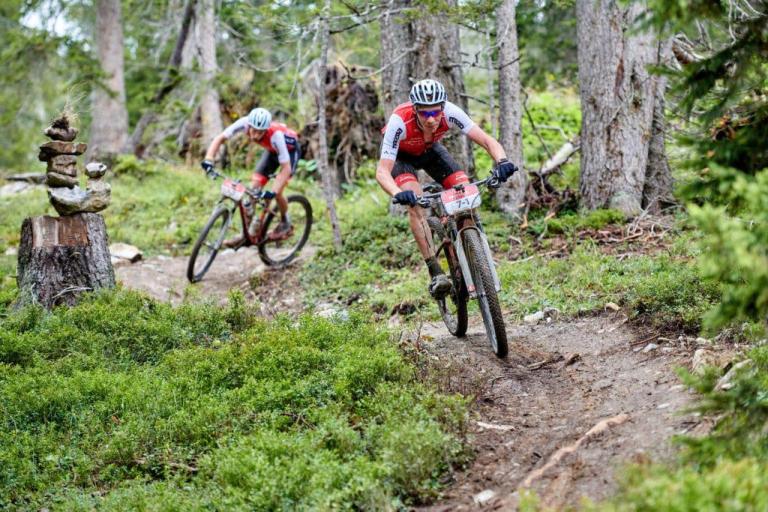 Two bikers traveling through a wooded area and passing by some stacked rocks