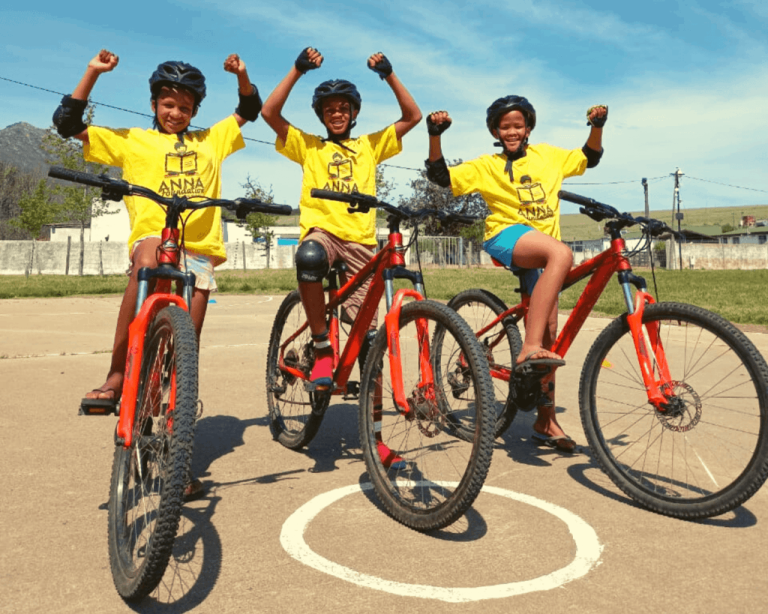 Three of the children from the anna foundation on their bikes hands in the air