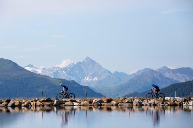 Two bikers with a lake in the foreground and mountain in the background
