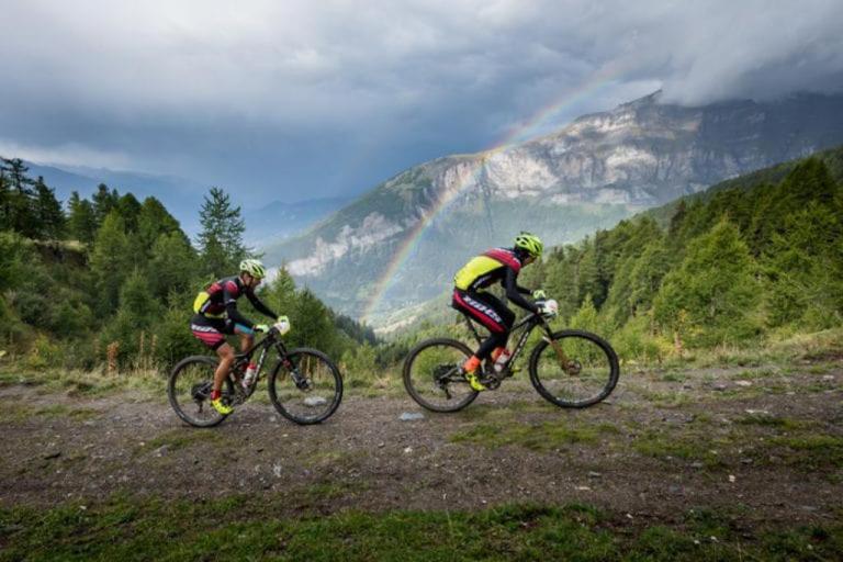 Two bikers peddling up a hill with a rainbow in the background