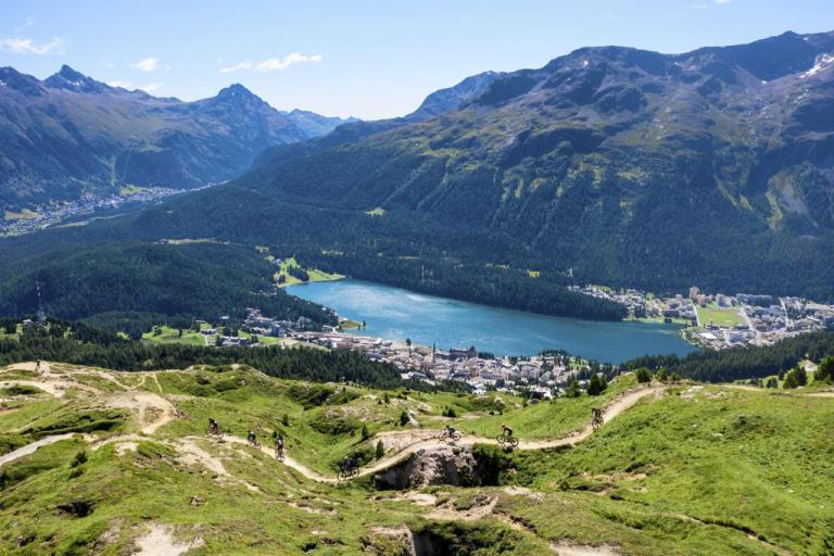 View of a lake nestled between mountains and riders riding a trail in Engadin at Swiss Epic