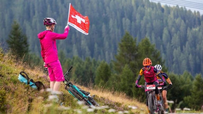 A man holding a Swiss flag while two bikers head past him [Swiss Epic]