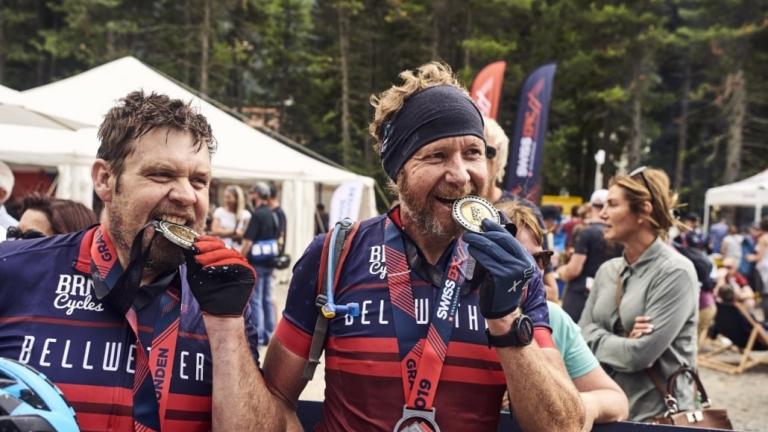 Two bikers biting their Swiss Epic Medals [Swiss Epic]