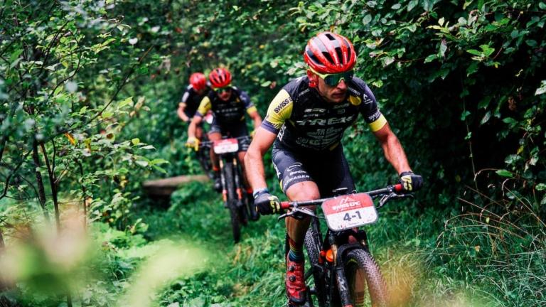 Three bikers wearing burnt orange helmets biking through a forest [Swiss Epic]