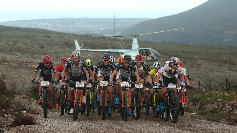 Bikers heading up a hill with a helicopter in the background [Absa Cape Epic]