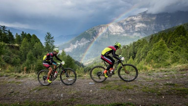 Two bikers peddling up a hill with a rainbow in the background [Swiss Epic]