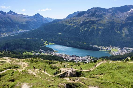 View of a lake nestled between mountains and riders riding a trail in Engadin at Swiss Epic