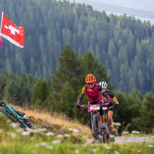 A man holding a Swiss flag while two bikers head past him [Swiss Epic]
