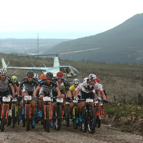 Bikers heading up a hill with a helicopter in the background [Absa Cape Epic]