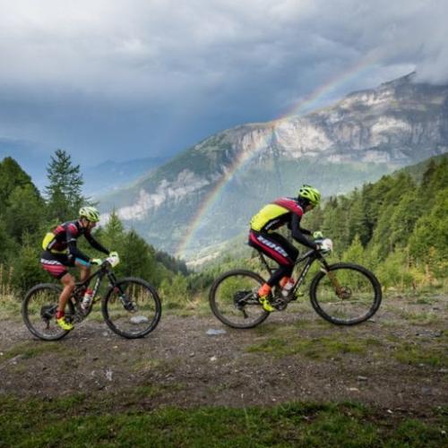 Two bikers peddling up a hill with a rainbow in the background [Swiss Epic]